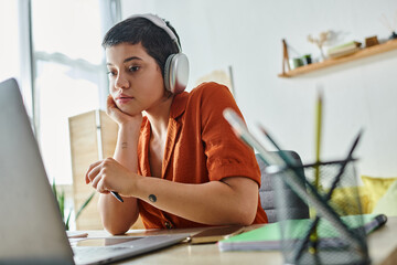 pensive young woman with tattoos holding pen and looking attentively at her laptop, studying