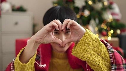 Wall Mural - Young chinese woman doing heart gesture sitting on sofa by christmas tree at home