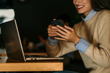 Wall Mural - Smiling Latina woman using a smartphone and working on a laptop in the bright cafe. Man in the background.