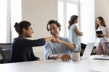 Two cheerful office friends celebrating business task completion, successful project startup, job promotion, sitting at shared workplace, giving fist bumps, talking, smiling, laughing