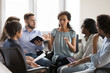 © fizkes - Serious African American group leader woman speaking on therapy meeting. Business team sitting on chairs in circle, talking, discussing solutions, project ideas, brainstorming