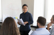 © fizkes - Happy confident Indian businesswoman presenting project to colleagues, talking to team of employees on corporate meeting, holding paper documents. Female mentor training group