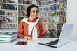 © Liubomir - Joyful curly Hispanic girl sitting at a table in front of a laptop in a headset looking at the camera and waving at the camera, office worker in a call center, working video call.