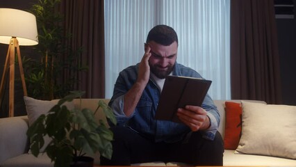 Wall Mural - Young bearded adult man sitting on sofa at home while using tablet having headache. Tired young man get having pain from a headache or migraine