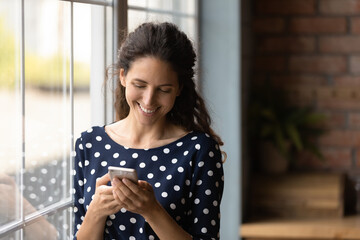 Wall Mural - Smiling woman using smartphone, standing near window at home, happy young female looking at phone screen, chatting with boyfriend in social network or shopping online, reading pleasant news