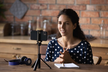 Wall Mural - Smiling woman recording video, using smartphone standing on tripod, sitting at wooden table at home, friendly young female teacher mentor shooting webinar or leading online lesson, explaining