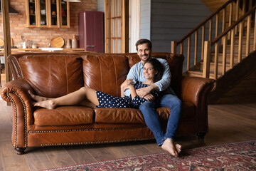 Wall Mural - Full length portrait happy millennial couple relaxing on cozy couch, smiling woman lying on man laps, looking at camera, pleasant family spending weekend in modern living room together, hugging