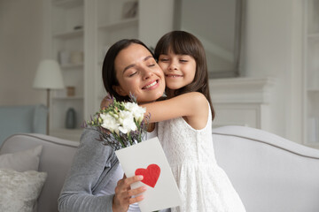 Wall Mural - Little daughter hugging happy young mother, congratulating with birthday or mothers day, smiling mother holding flowers and greeting card with red heart, sitting together on cozy sofa at home