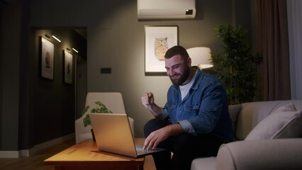 Bearded young adult man sitting on sofa while using laptop checking messages read notice receive good news on e-mail celebrate great opportunity feels happy. Special offer, student got scholarship