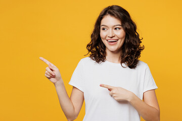 Wall Mural - Young smiling happy Caucasian woman she wear white blank t-shirt casual clothes point index finger aside on area mock up isolated on plain yellow orange background studio portrait. Lifestyle concept.