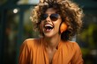 © Georgii - Cheerful African American girl in orange dress enjoying favorite playlist in headphones. Happy smiling young woman listening to music or podcast in app, having fun and laughing. Blurred background.