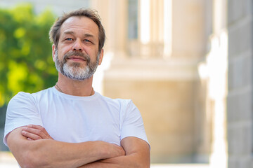 Wall Mural - A handsome middle-aged man with a beard stands on a blurred city background, crossing his arms and smiling in a relaxed and joyful manner. success and optimism.