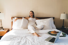 Cheerful woman resting with laptop and breakfast tray on bed