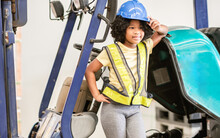 Little Girl Wearing Hard Hat, Driving Tractor In Factory, Smiling, Posing As Worker With Happiness, Doing Activities On Weekend. Industry, Education Concept.