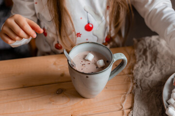 cute little girl in Santa hat and pajamas drinks cocoa with marshmallows in the kitchen at home on Christmas day