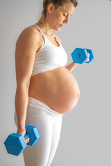 Wall Mural - Side shot of pregnant mother with round tummy  in white sports wear and working out with arm weights. Last month of pregnancy - week 39. White background. Bright shot.