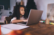© BullRun - Thoughtful ethnic girl sitting at wooden table while working on laptop