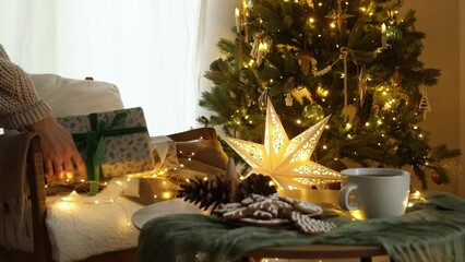 Wall Mural - Merry Christmas! Hands in cozy sweater putting stylish wrapped gift on modern chair at festive christmas tree with glowing lights in cozy room. Atmospheric christmas eve footage