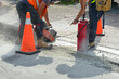 Two construction workers wearing belts cut a hole in a concrete sidewalk using a mechanical saw. An orange safety cone is on the road and sidewalk. Concrete dust is blowing around the street. 

