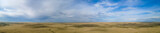 Fototapeta  - Aerial panorama of a vast flat dry prairie landscape with no trees under a sky with white to blueish colored clouds and patches of clear bright blue sky.
