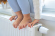 Hands and bare feet of child on heater. Child sitting by window and warming up from the heating radiator. Heating in an apartment, at home.