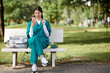 © DragonImages - Cheerful medical nurse sitting on bench in park, eating lunch and scrolling through social media