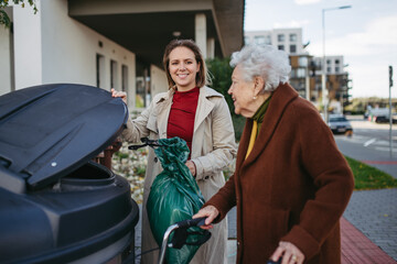 woman helping elderly neighbor throw away trash into garbage can, waste container in front of their 