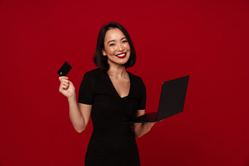 Wall Mural - Joyful woman using laptop computer and credit card for online shopping isolated over red wall