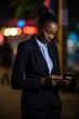 © Stavros - African American businesswoman in a formal suit with a smartphone. On the background of the street of the business district of the night city. Online monitoring of stock reports.