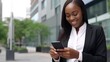 © Stavros's son - Cheerful young African American businesswoman in a formal suit with a smartphone. On the background of the street of the business district of the city. Online monitoring of stock reports.