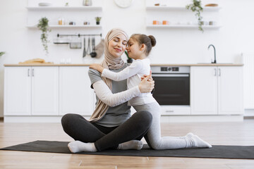 Wall Mural - Adorable girl in sport outfit standing on knees on mat while trading positive energy with mom during exercises. Loving muslim female in hijab hugging daughter during fitness routine at home.