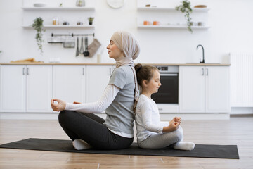 Wall Mural - Energetic little girl sitting back to back with muslim lady during her achieving cross-legged position. Happy mother promoting daughter's sense of calmness by meditating together at home.