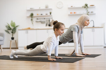 Wall Mural - Attractive muslim mother in hijab practicing plank position with baby daughter in kitchen after wake up in morning. Feeling so comfortable and relax. Yoga of woman with her baby girl for healthcare.