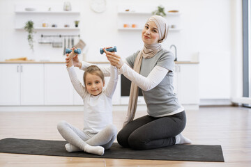 Wall Mural - Arabian family, woman in hijab helping sweet daughter with lifting dumbbells on black mat in kitchen. Cheerful female parent encouraging healthy habits while enhancing kid's strength at home.