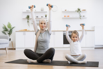 Wall Mural - Emotional muslim woman and kid in sports clothes raising weights with excitement while sitting on room floor. Happy young mom and baby girl exercising together with joy on mat at home.