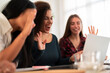 © Studio Marmellata - Group of happy young coworkers sitting at table with laptop while waving hands during video call meeting with colleague in modern workspace at daylight
