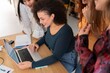 © Studio Marmellata - Group of cheerful multiracial female colleagues in casual clothes smiling and looking at screen while gathered at table with laptop and working together on project in modern workspace