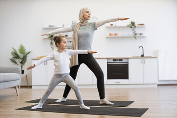 Wall Mural - Focused muslim woman with her cute daughter in sport clothes standing at cozy kitchen and practising yoga exercise on mat. Concept of concentration, relaxation and workout.