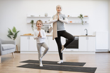 Wall Mural - Full length of muslim lady in hijab balancing in standing asana with cute girl in sporty outfit on yoga mats. Relaxed woman calming mind while sweet daughter trading positive emotions from mom indoors