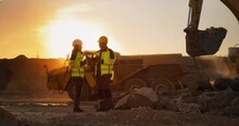 Cinematic Golden Hour Shot Of Construction Site: Caucasian Male Civil Engineer And Hispanic Female Urban Planner Talking, Using Tablet. Trucks, Excavators, Loaders Working To Build New Apartment Block