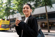 © Andrei - Attractive multiracial young businesswoman with a cheerful expression, waiting in the street for the bus to the office, drinking her morning coffee