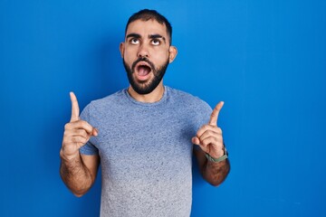 Poster - Middle east man with beard standing over blue background amazed and surprised looking up and pointing with fingers and raised arms.