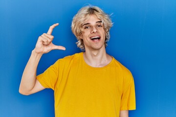 Poster - Young modern man standing over blue background smiling and confident gesturing with hand doing small size sign with fingers looking and the camera. measure concept.