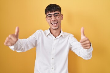 Young hispanic man standing over yellow background approving doing positive gesture with hand, thumbs up smiling and happy for success. winner gesture.