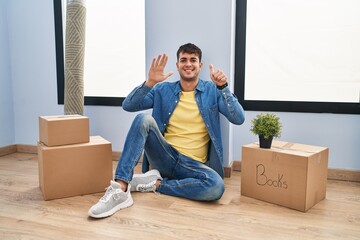 Wall Mural - Young hispanic man sitting on the floor at new home showing and pointing up with fingers number six while smiling confident and happy.