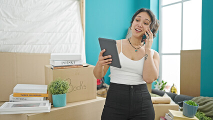 Canvas Print - Young beautiful hispanic woman using touchpad talking on smartphone at new home