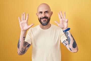 Wall Mural - Hispanic man with tattoos standing over yellow background showing and pointing up with fingers number nine while smiling confident and happy.