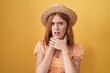Young redhead woman standing over yellow background wearing summer hat shouting and suffocate because painful strangle. health problem. asphyxiate and suicide concept.