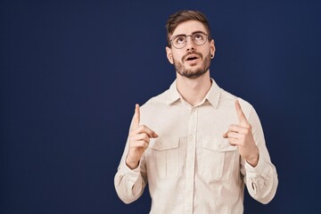 Poster - Hispanic man with beard standing over blue background amazed and surprised looking up and pointing with fingers and raised arms.