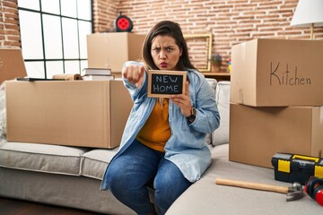 Wall Mural - Young hispanic woman holding blackboard with new home text pointing with finger to the camera and to you, confident gesture looking serious